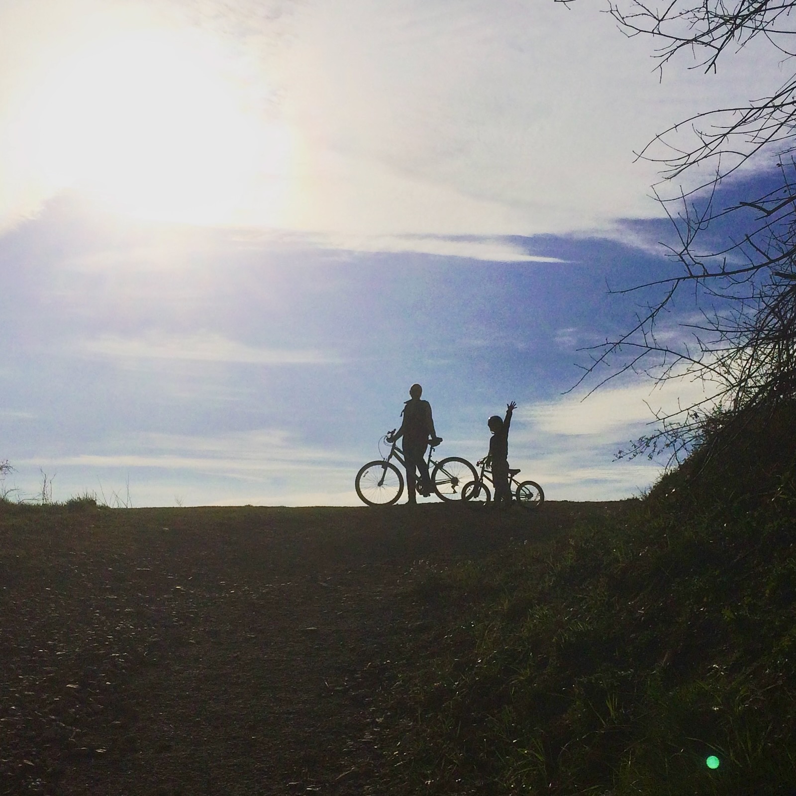 Silhouettes d’un adulte et d’un enfant faisant une pause à vélo sur un chemin en pleine nature sous un ciel ensoleillé, près de L’Autre Maison.
